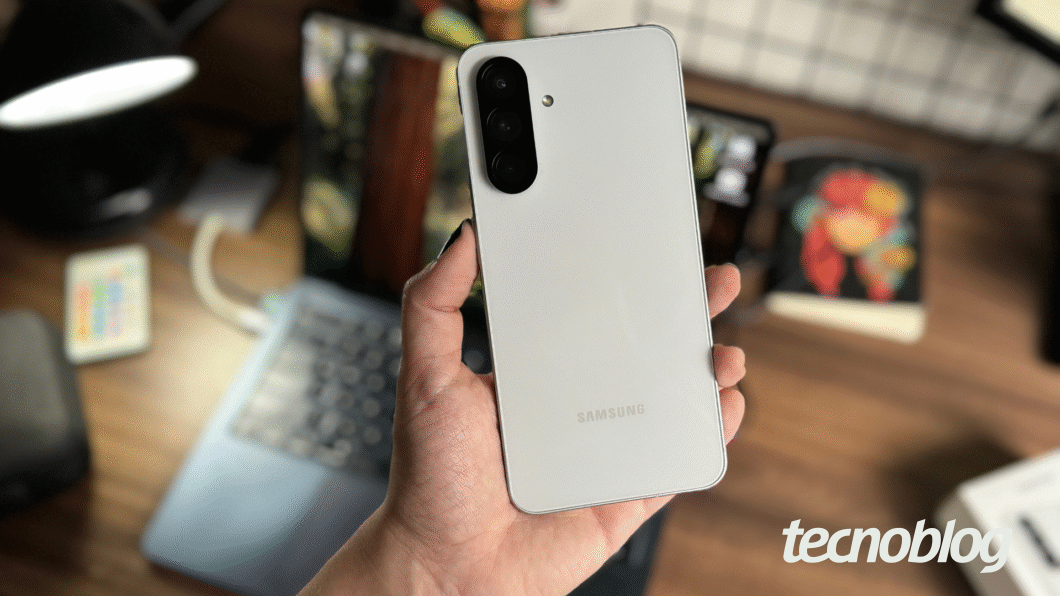 Imagem mostra mão segurando celular Samsung Galaxy A56, exibindo a parte de trás do aparelho na cor cinza; ao fundo, mesa de trabalho desfocada