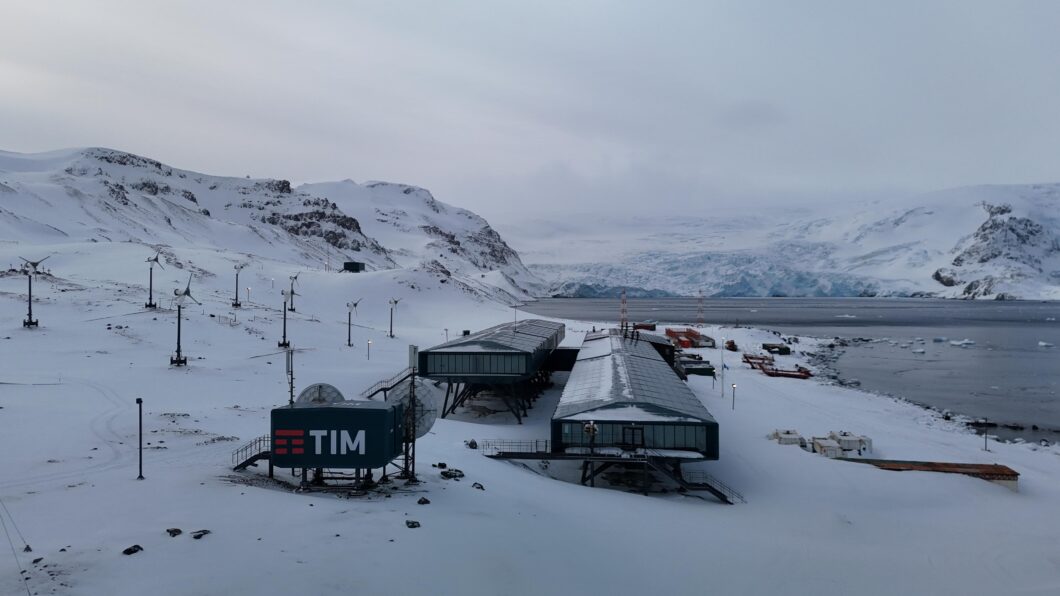 Vista panorâmica da Estação Antártica Comandante Ferraz (Brasil) coberta de neve, localizada na Baía do Almirantado. A imagem mostra a moderna estrutura principal da estação em plano elevado, com grandes janelas, e o módulo de comunicação da TIM em primeiro plano (um edifício preto com o logo da TIM em branco e vermelho). Diversas turbinas eólicas e antenas de satélite cercam a estação, em contraste com as montanhas e geleiras ao fundo, e a água gelada do oceano.