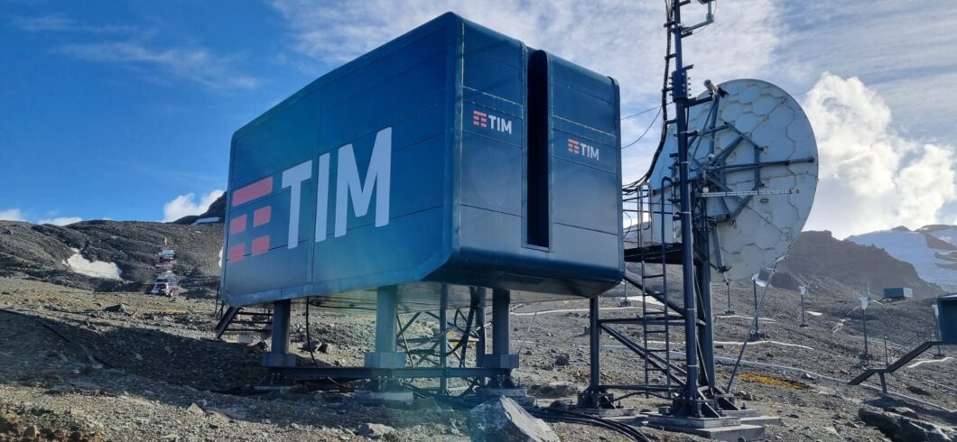 Close-up de um módulo de telecomunicações da TIM na Antártica, provavelmente na área da Estação Comandante Ferraz. O módulo é um grande contêiner elevado, na cor azul-escura, com o logo da TIM em destaque. Ao lado, há uma grande antena parabólica e estruturas de comunicação em metal. O céu está azul e parcialmente ensolarado, e o solo é rochoso e árido, com manchas de neve e gelo.