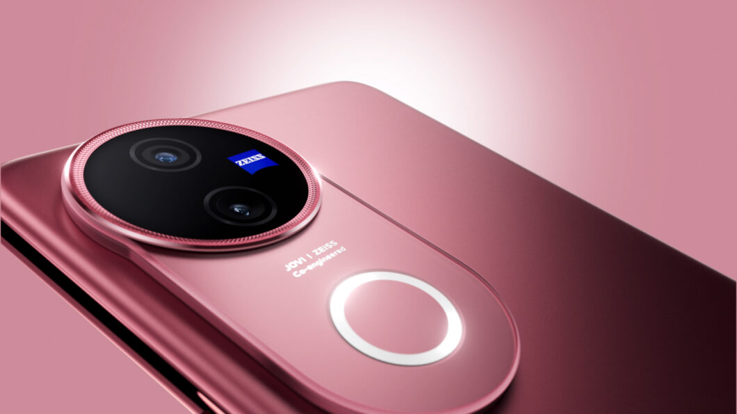 Close-up da parte traseira de um smartphone dobrável na cor rosa metálico. A imagem destaca o módulo de câmeras circulares duplas em uma base preta, cercada por uma borda texturizada da mesma cor do aparelho. No centro da base preta há um pequeno logotipo azul com a palavra "ZEISS" em branco. Abaixo, estão as inscrições "JOVI | ZEISS Co-engineered" e um anel metálico circular liso. Fundo em tom rosa degradê.