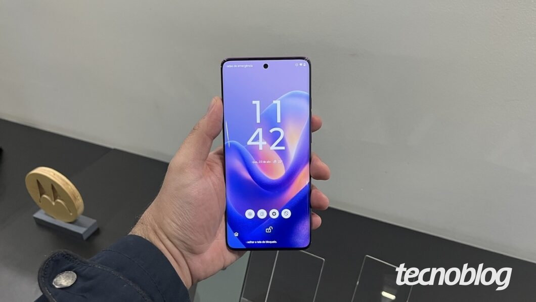A imagem mostra o Motorola Edge 60 sendo segurado com uma mão. A tela do dispositivo está visível, exibindo a hora 11:42, com um fundo em tons de azul e roxo. O sistema operacional parece estar com a interface personalizada da Motorola. No fundo, há um logo da Motorola em um suporte de exposição.
