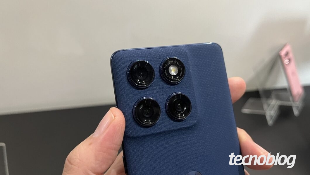 A imagem mostra a parte traseira do Motorola Edge 60, com foco na área das câmeras. O dispositivo possui um módulo de câmeras quadrado, com três lentes circulares e um flash LED. O aparelho é de cor azul escuro e está sendo segurado com uma mão. Ao fundo, é possível ver um suporte transparente com outros aparelhos.