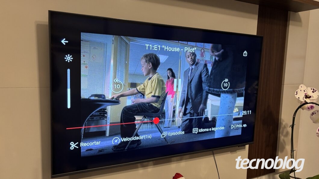 Alt text: Televisor exibe uma cena do episódio “T1:E1 ‘House – Pilot’”, mostrando um menino sentado em uma cadeira baixa enquanto adultos observam ao fundo. Na tela aparecem controles do player, como barra de progresso, botões de retroceder e avançar “10”, pausa, “Velocidade (1×)”, “Episódios”, “Idioma e legendas” e “Próx. ep.”. À direita da TV há uma orquídea branca em um vaso.