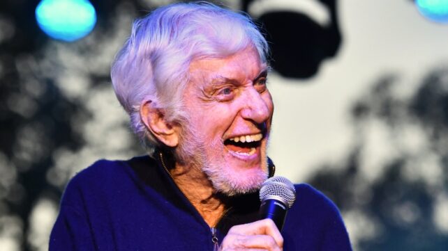 A grinning Dick Van Dyke speaking into a microphone on stage