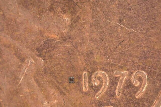 Close-up image of a tiny black square positioned to the left of the numbers '1979' on a copper-colored penny, which has scratch marks from wear.