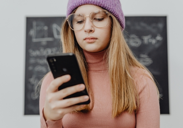 girl with glasses and purple beanie looking at her smart phone with a blackboard in the background
