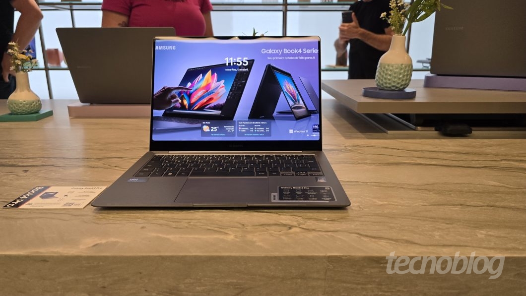 Notebook da série Galaxy Book4 da Samsung, aberto, em cima de uma mesa de mármore, acompanhado de jarros com flores, pessoas ao fundo e outros notebooks da linha desfocados