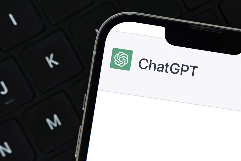 Close-up da tela de um smartphone exibindo o logo e o nome do aplicativo ChatGPT, com um teclado de computador desfocado ao fundo.