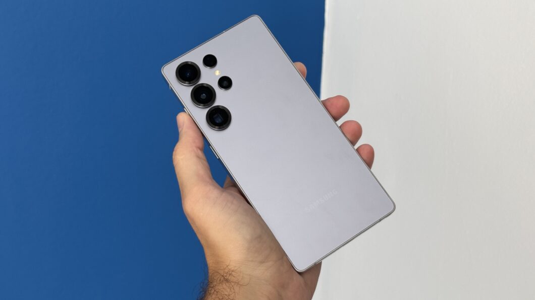 Uma mão segurando o Galaxy S25 Ultra com a traseira equipada com câmeras virada para frente. Ao fundo paredes na cor azul e branco.