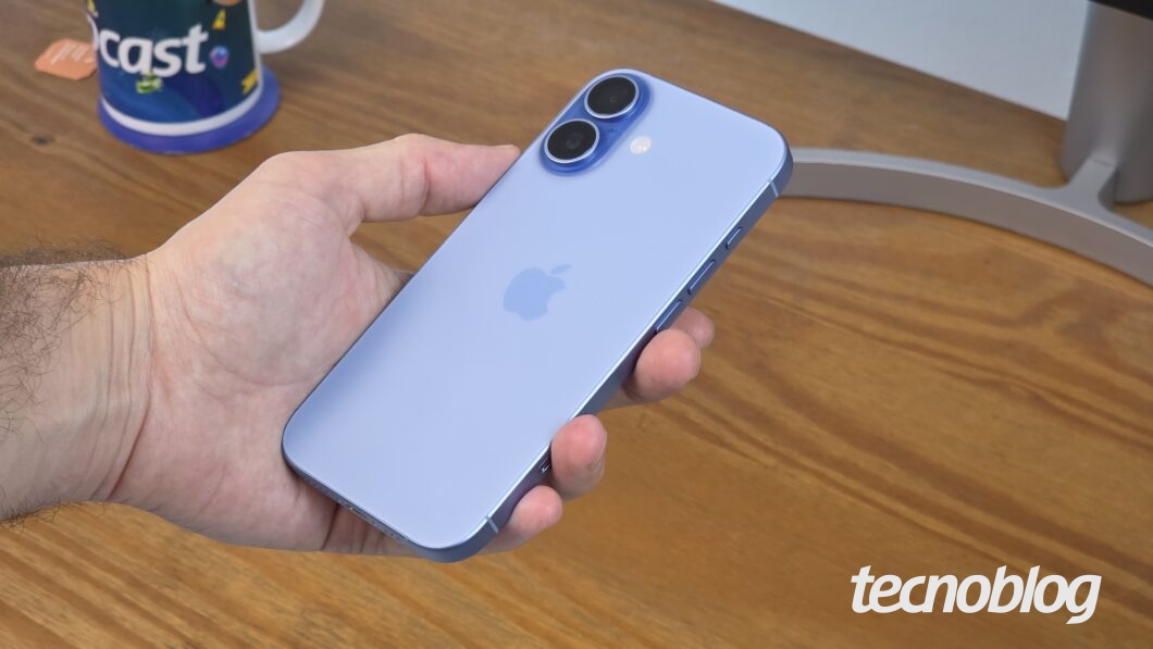 Mão segurando iPhone 17 azul-névoa, com mesa em madeira como fundo