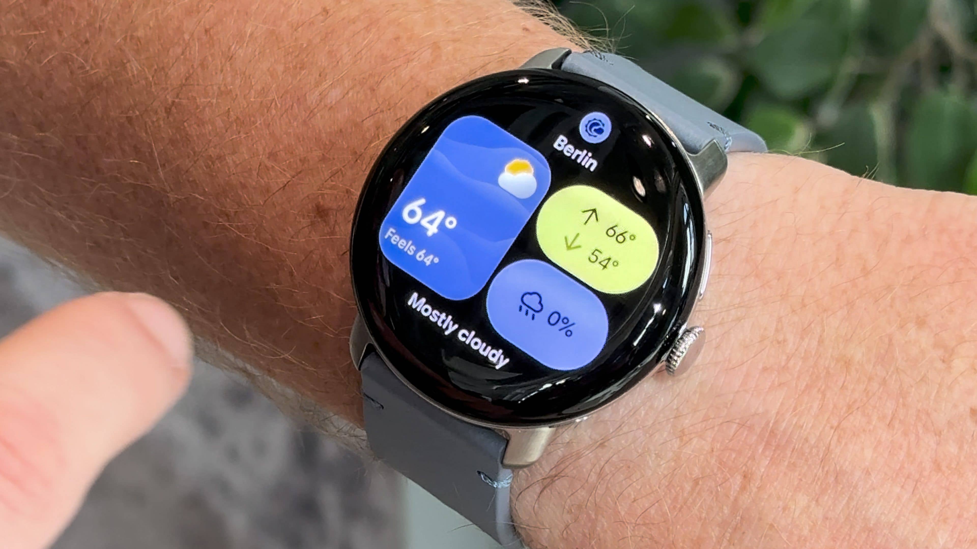 A close-up of a Google Pixel Watch 4 displaying weather information for Berlin, showing 64°F and mostly cloudy.