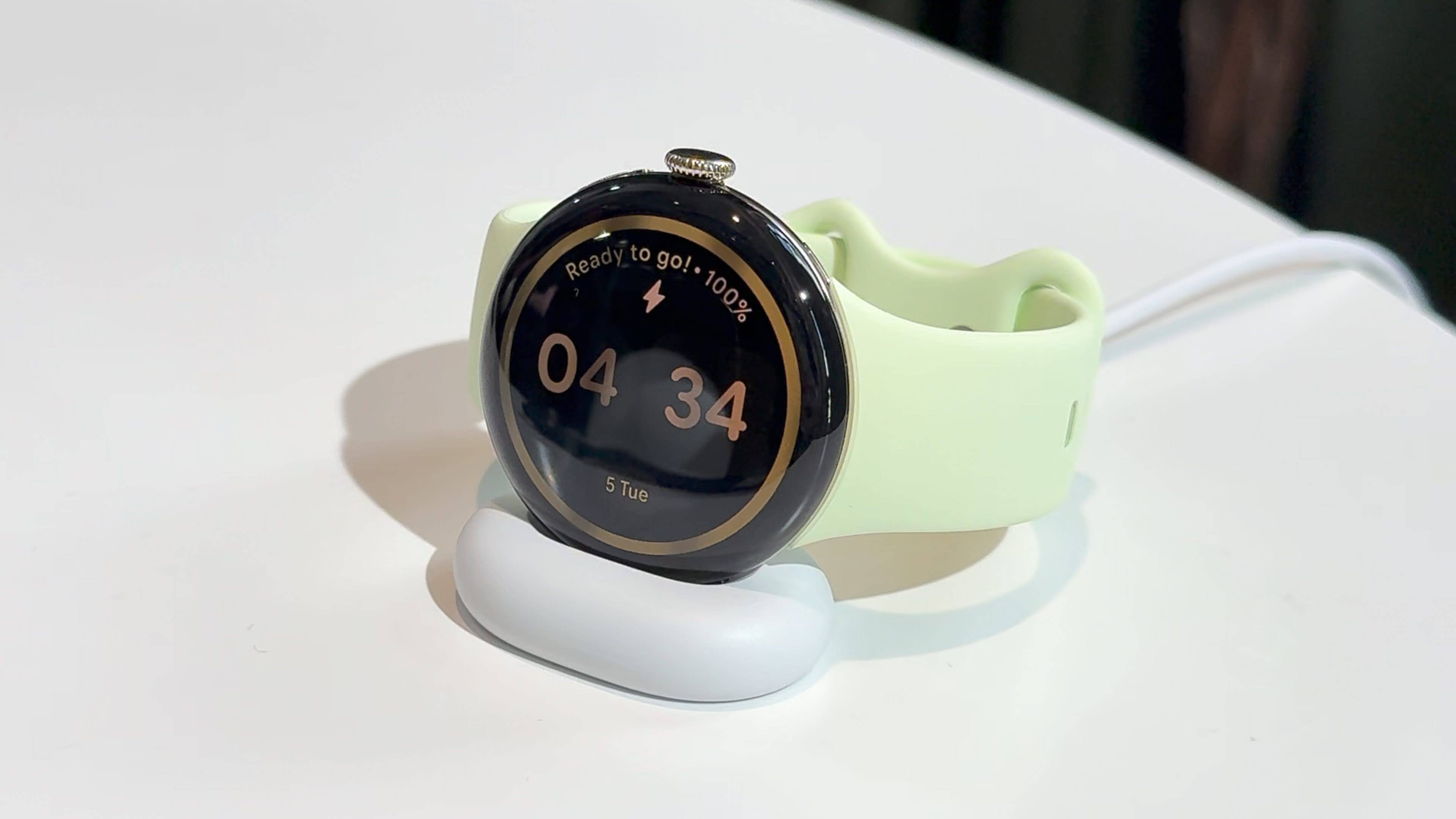 A Google Pixel Watch 4 on a charging dock, displaying time 04:34 and a battery status of 100%.