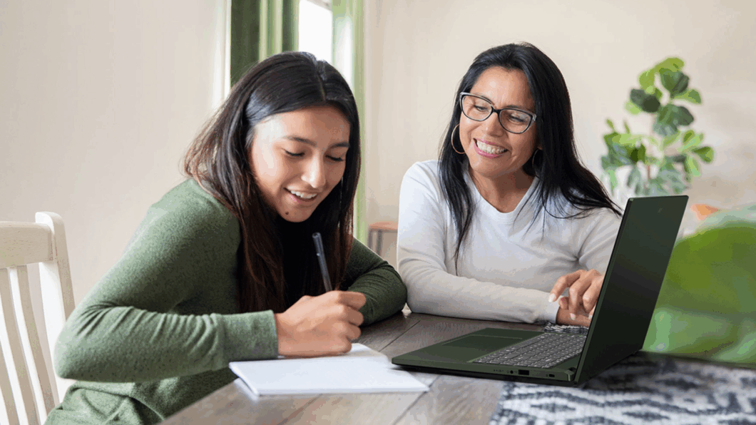 Imagem mostra duas mulheres sentadas a uma mesa com o notebook Acer Inspire GO 15 apoiado sobre ela. A mulher da esquerda escreve em uma folha de papel, enquanto a mulher da direita sorri e olha pra ela.