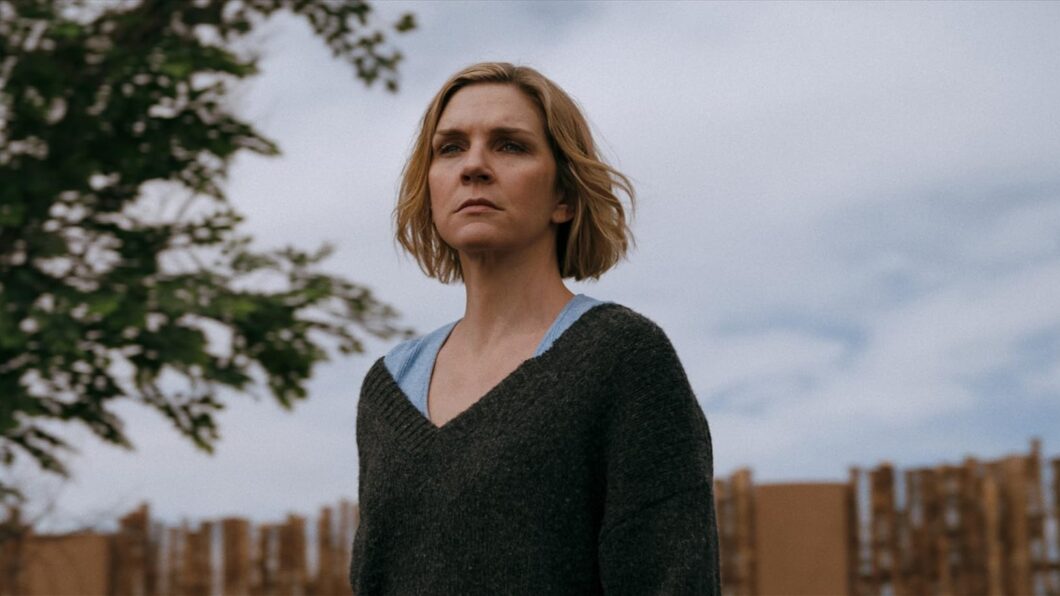 Imagem da série Pluribus, com a protagonista, Carol, uma mulher branca de cabelo curto e loiro, usa uma blusa preta. Ela está ao ar livre, o céu azul com nuvens ao fundo, uma cerca de madeira marrom na parte inferior e folhas de uma árvore aparecem no lado esquerdo da imagem.