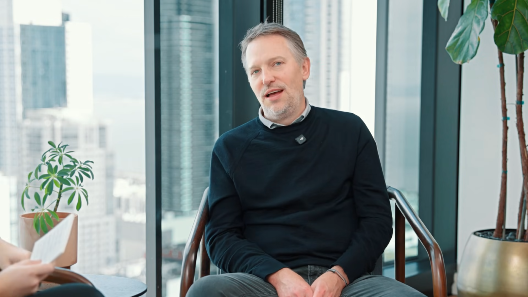 Homem branco de meia-idade, com cabelos grisalhos curtos, sentado em uma cadeira dentro de um escritório moderno. Ele veste blusa preta de mangas longas e calça cinza, fala enquanto olha para alguém fora do enquadramento. Ao fundo, janelas de vidro mostram prédios altos da cidade; há plantas em vasos ao redor, reforçando o ambiente corporativo.