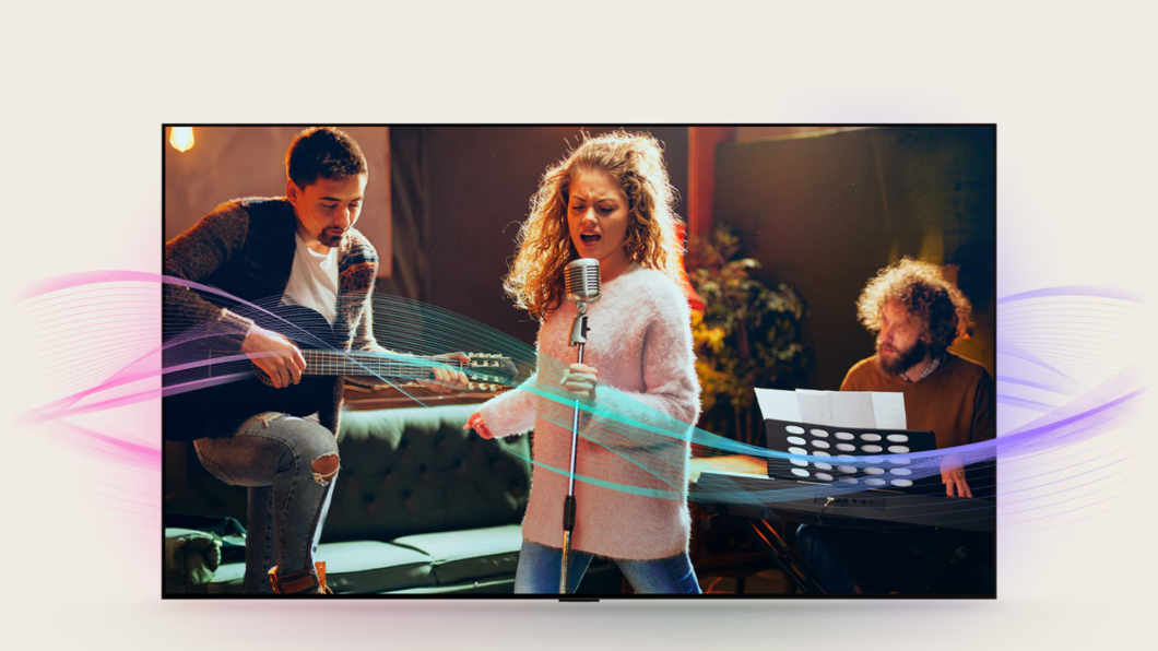 Mulher ao centro com microfone cantando, homem ao seu lado esquerdo com violão e outro homem a direita sentado.