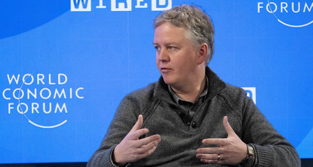 Imagem colorida mostra Matthew Prince, CEO da Cloudflare. Ele é um homem de cabelos grisalhos curtos, está posicionado no centro da imagem, falando e gesticulando com as mãos abertas. Ele veste um suéter cinza escuro sobre uma camisa preta. O fundo é uma tela azul vibrante com as palavras "WORLD ECONOMIC FORUM" impressas em letras brancas grandes à esquerda.