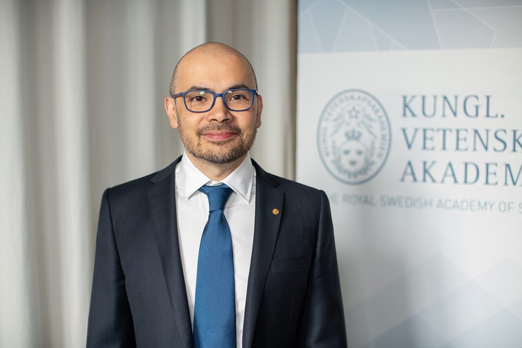 Foto de Demis Hassabis ao lado de uma placa branca. Ele veste um blazer preto, com uma gravata azul e uma camisa branca. 