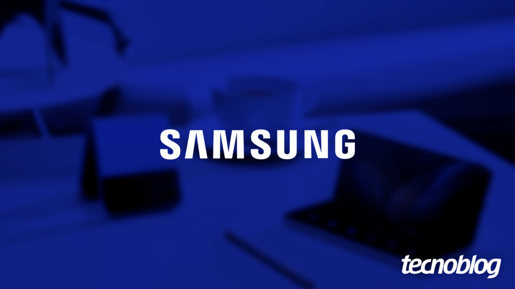 Imagem mostra a palavra "SAMSUNG" sendo exibida no centro, em letras brancas e maiúsculas. O fundo, em tom azul escuro, mostra elementos desfocados que sugerem um ambiente de escritório. Na parte inferior direita, o logotipo do "Tecnoblog" é visível.