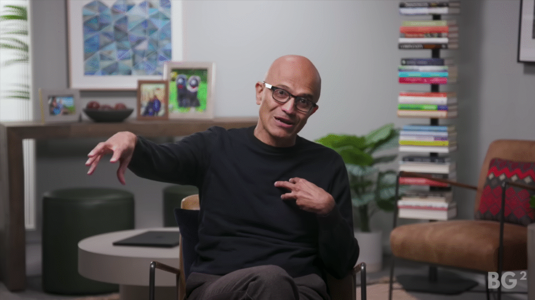 Homem calvo de óculos e suéter preto gesticula com a mão esquerda enquanto fala, sentado em uma cadeira em ambiente moderno e aconchegante, com livros empilhados verticalmente ao fundo, plantas e fotos emolduradas sobre uma mesa (imagem: reprodução/YouTube)