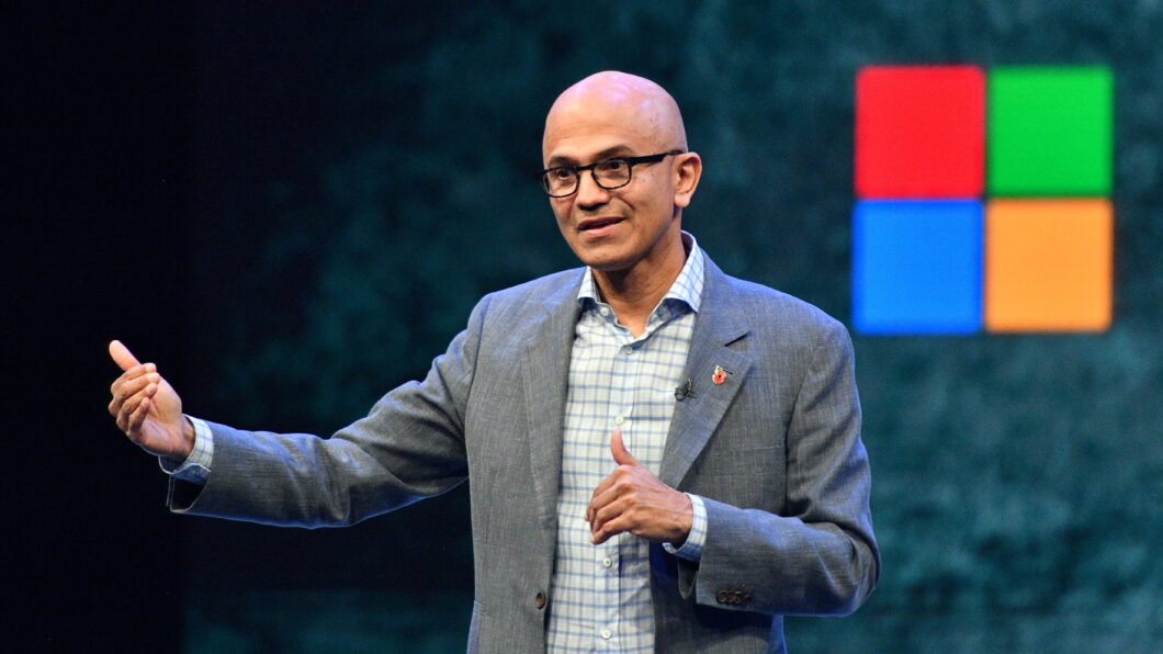 Satya Nadella, homem de óculos usando uma camisa cinza e um paletó cinza escuro. Ao lado, um logo do Windows.