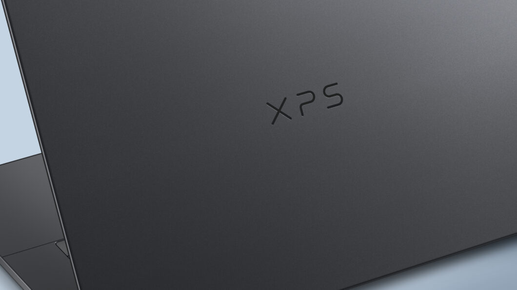 Parte traseira de um notebook fechado, vista em close, com acabamento escuro e fosco. No centro da tampa aparece o logotipo “XPS” gravado de forma discreta. À esquerda, é possível ver parcialmente a dobradiça e a borda do equipamento, sugerindo um design fino e minimalista.