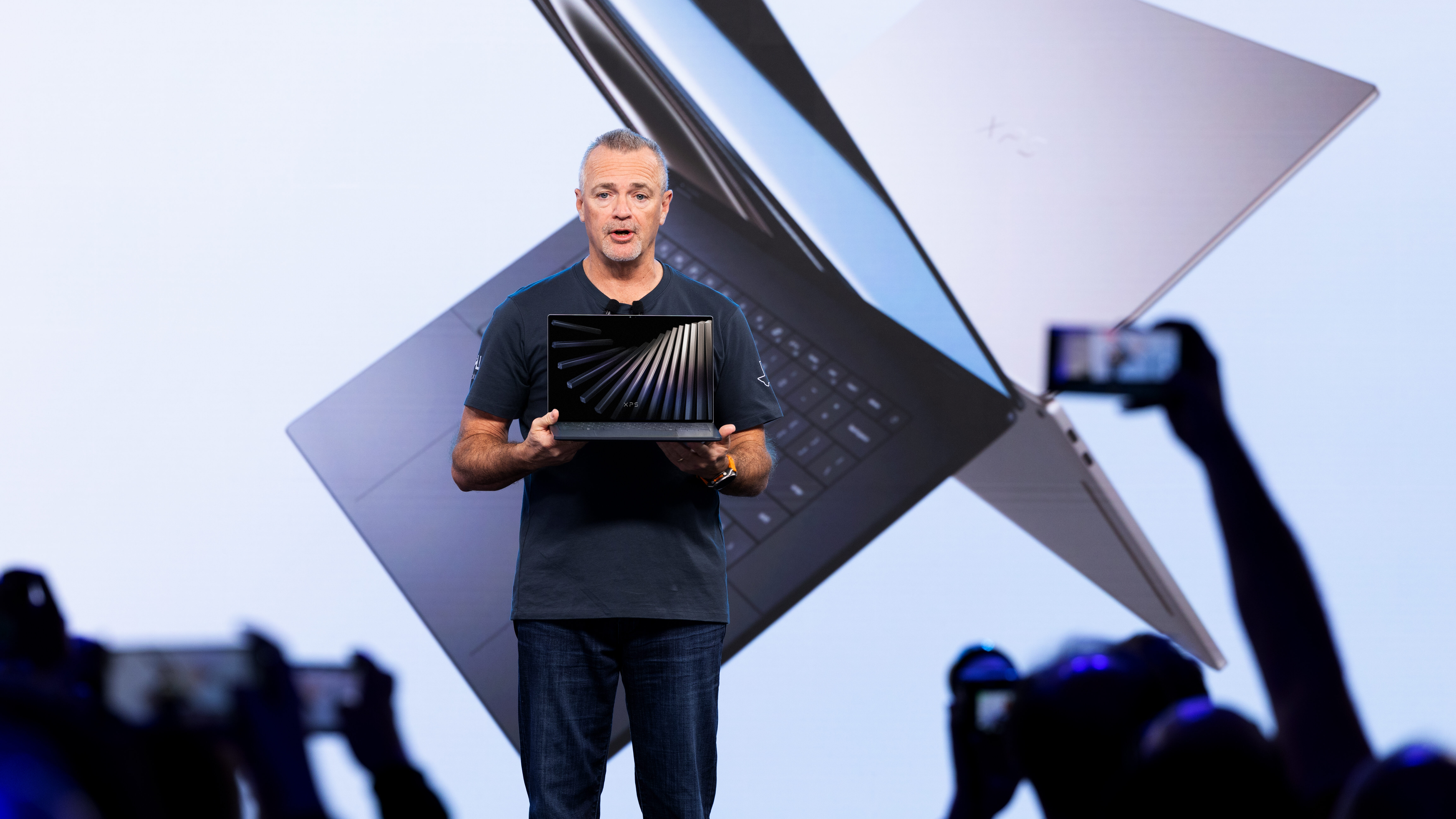 Fotografia colorida mostra o executivo Jeff Clarke no centro de um palco, segurando um notebook aberto. Ele é um homem branco de cabelos curtos grisalhos e veste camiseta azul-marinho e calça jeans. Ao fundo, um telão exibe a imagem ampliada de dois notebooks prata que se cruzam, com a inscrição "XPS" visível em um deles. Em primeiro plano, silhuetas de pessoas na plateia levantam celulares para fotografar o momento. O ambiente tem iluminação clara e tons azulados.