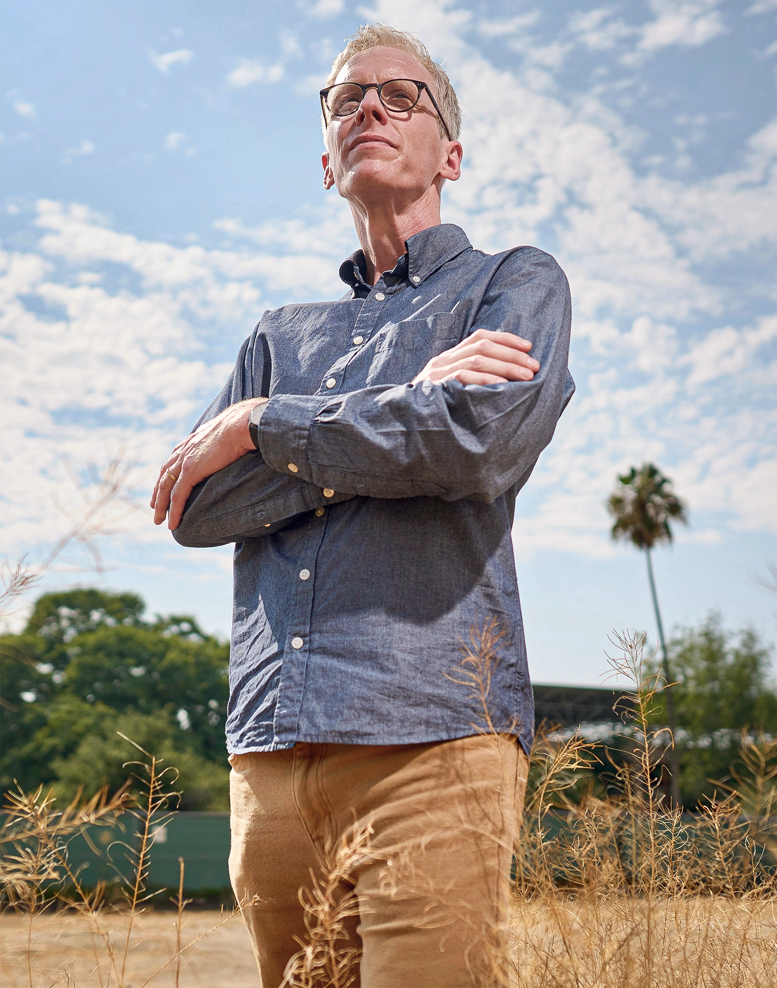 A man wearing glasses and a blue shirt stands in a field under a cloudy sky.