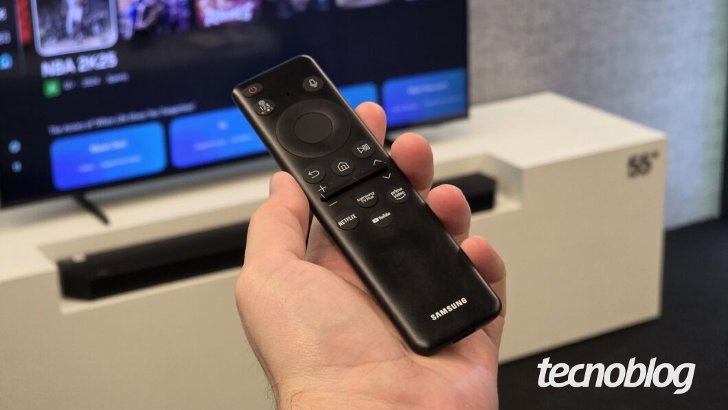 Mão segura controle remoto preto Samsung com botões de voz e acesso rápido a Netflix, YouTube e Prime Video, TV ao fundo.