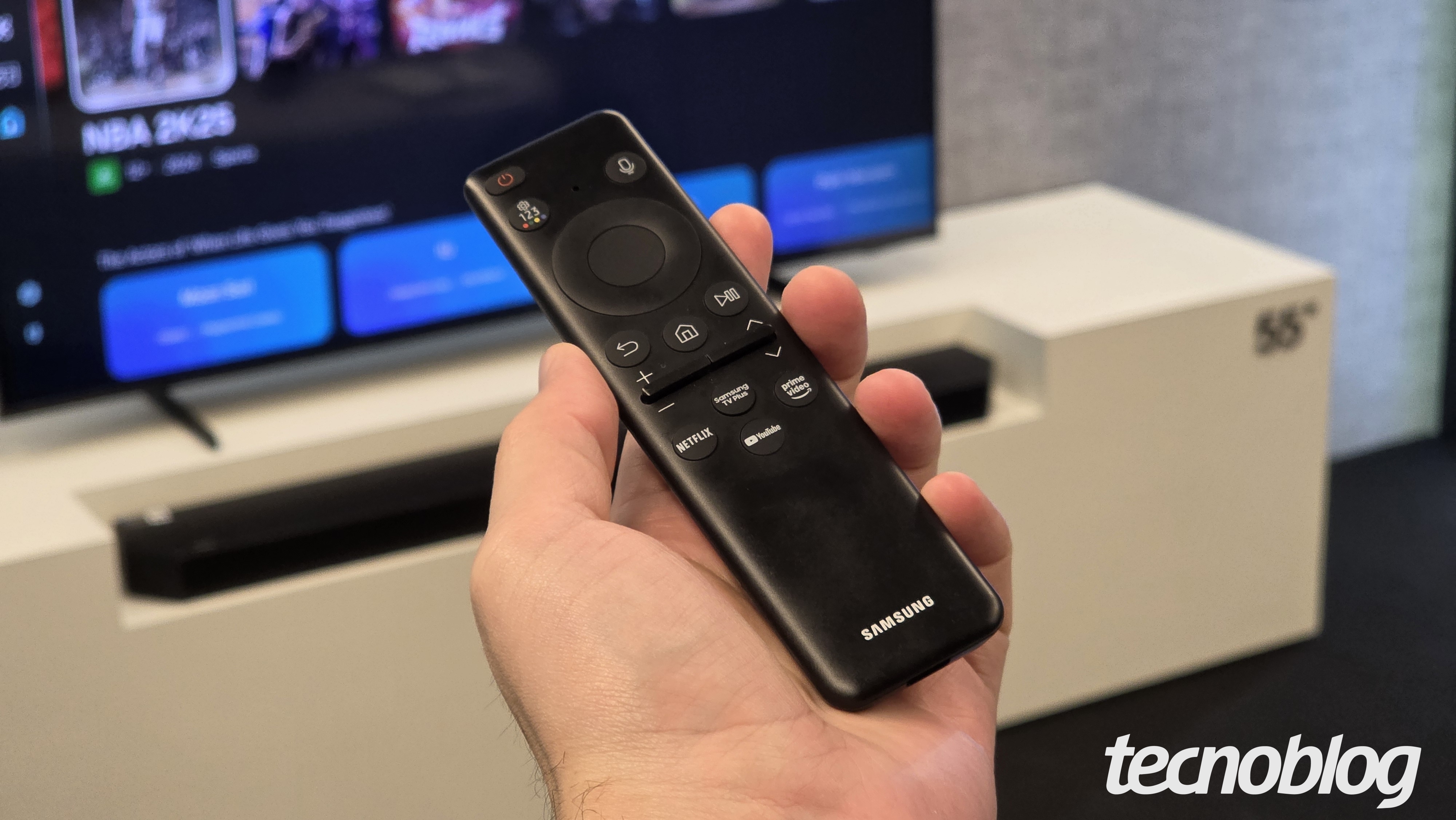 Mão segura controle remoto preto Samsung com botões de voz e acesso rápido a Netflix, YouTube e Prime Video, TV ao fundo.