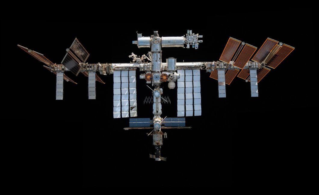 Fotografia da Estação Espacial Internacional (ISS) centralizada contra o fundo totalmente preto do espaço. A estrutura metálica complexa apresenta diversos módulos cilíndricos, braços robóticos e grandes painéis solares retangulares dispostos simetricamente nas extremidades. Os painéis à direita possuem tons alaranjados e bronze, enquanto os da esquerda e do centro exibem tons prateados, cinzas e azulados. A iluminação é lateral, destacando as texturas metálicas e as sombras da engenharia espacial.