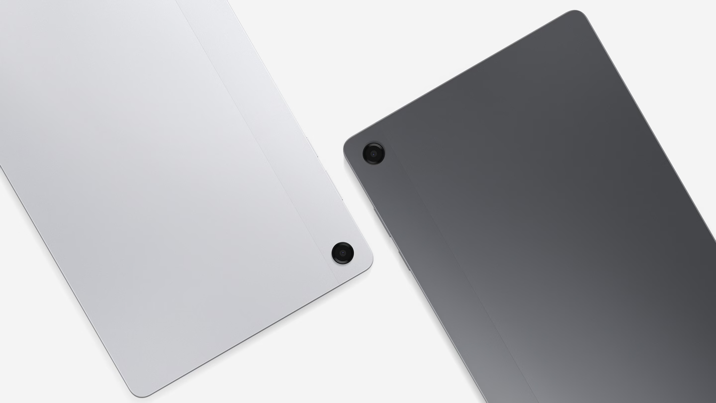 Close-up da parte traseira de dois tablets Galaxy Tab A11 Plus dispostos diagonalmente. À esquerda, o modelo na cor prata e, à direita, o modelo em grafite escuro. O foco destaca o acabamento metálico e a lente única da câmera traseira, que é circular e levemente saltada, posicionada no canto superior. Uma linha vertical sutil atravessa a parte superior da carcaça, separando a área da antena do restante do corpo do dispositivo.