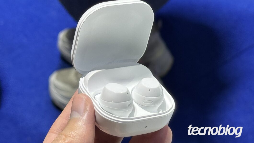 Uma mão segura um estojo de fones de ouvido sem fio de cor branca, que está aberto e revela dois fones também brancos. O logotipo "tecnoblog" está no canto inferior direito, em branco.