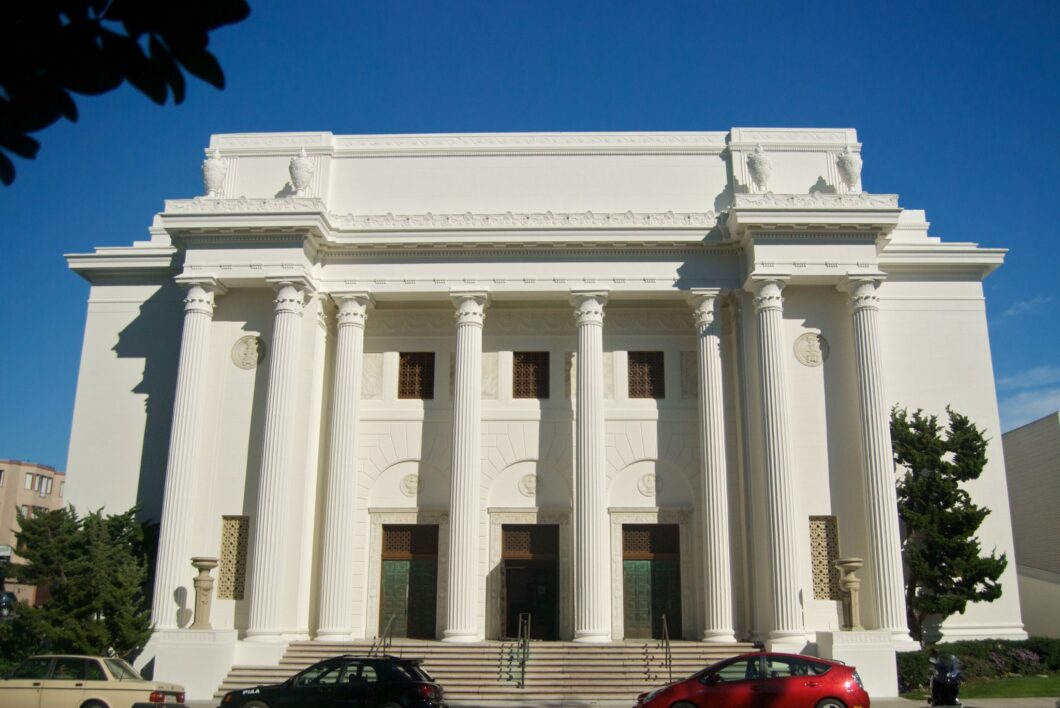 Fotografia frontal de um edifício neoclássico branco sob céu azul límpido. A fachada apresenta oito colunas monumentais de estilo jônico que sustentam um frontão reto adornado com quatro grandes vasos ornamentais no topo. No centro, três portas altas de cor esverdeada são precedidas por uma escadaria de mármore. Acima das portas, há janelas quadradas com treliças escuras. Árvores verdes flanqueiam o prédio, e na base da imagem, três carros — um bege, um preto e um vermelho — estão estacionados na rua.