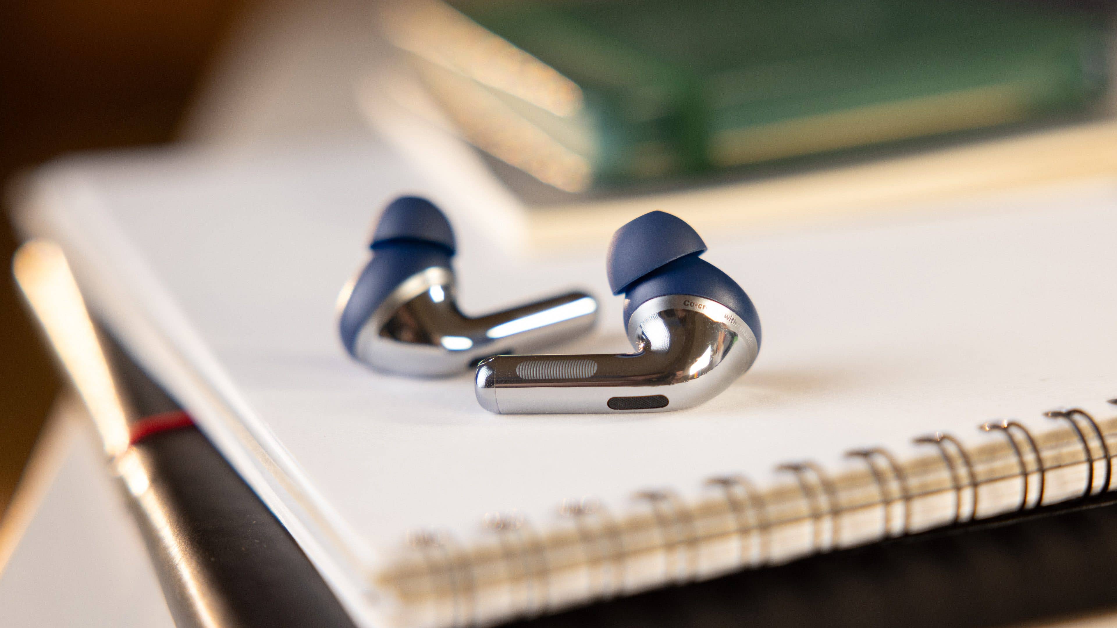 The OnePlus Buds Pro 3 wireless earbuds on a notebook with pens and books in the background.