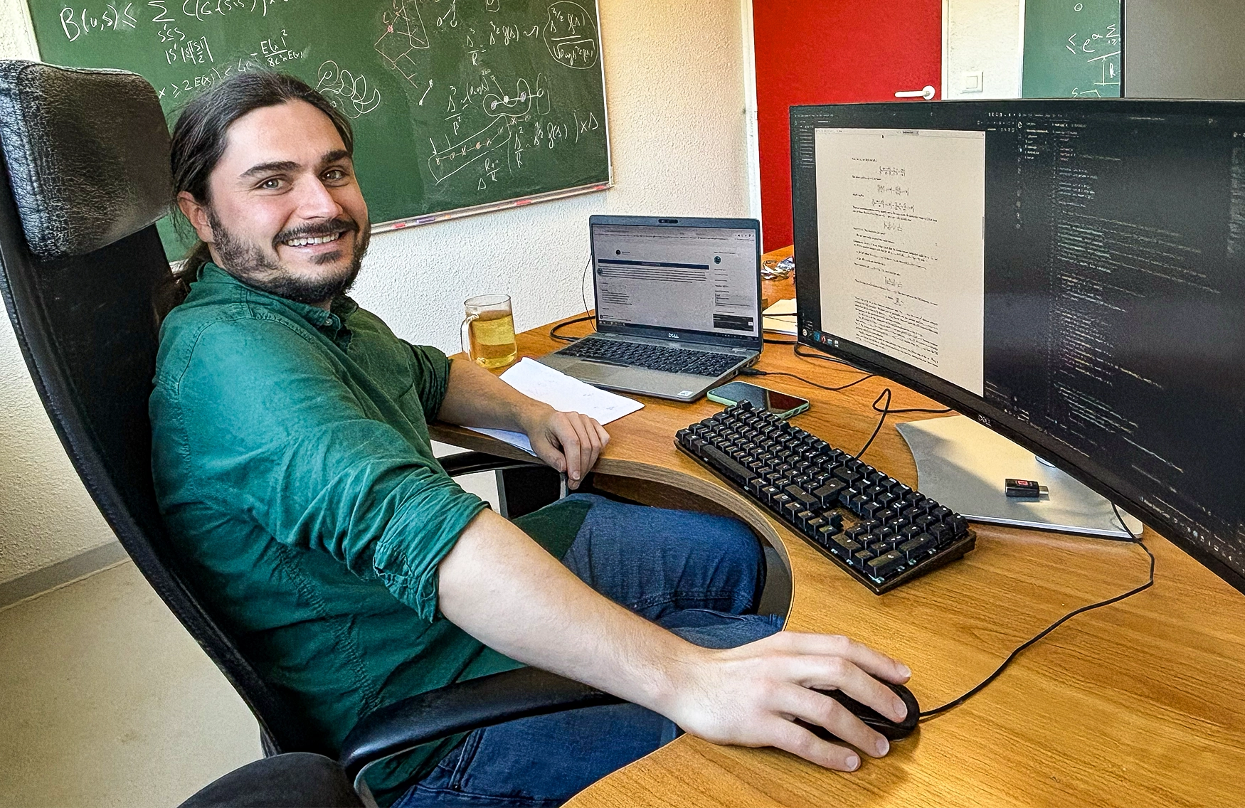 Man wearing a green shirt, smiling and writing on a blackboard.