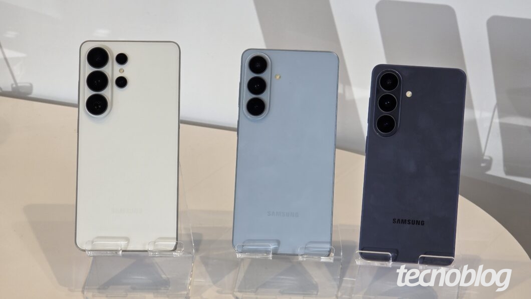 Galaxy S26 Ultra, S26 Plus e S26 vistos de costas. Os aparelhos estão sobre suportes transparentes. O S26 Ultra é o maior deles e tem cor dourada. O S26 Plus é o intermediário, um pouco menor que o Ultra, e está na cor cinza. O S26 é o menor dos três e está na cor preta.