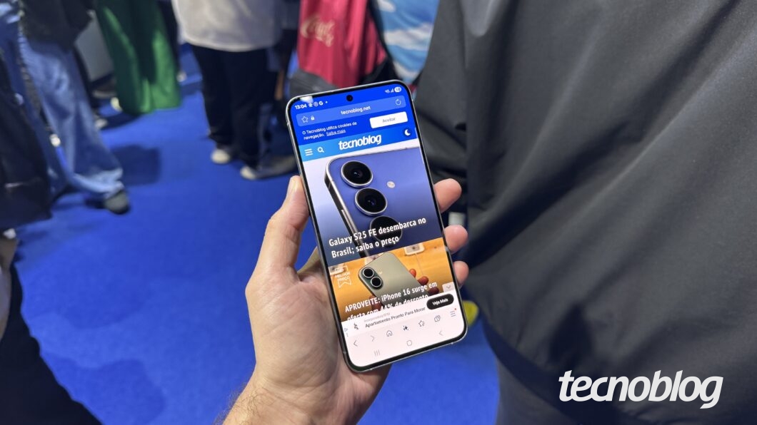 Galaxy S25 FE segurado por uma mão com a tela pra cima e mostrando a home page do Tecnoblog.