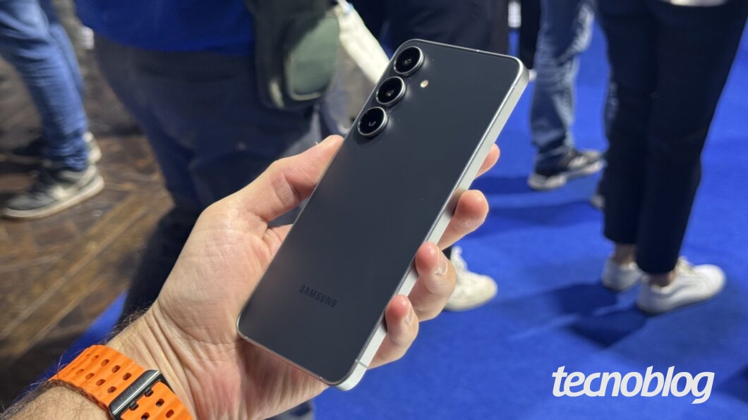 Uma mão segura um smartphone Galaxy S25 FE preto e prata, exibindo a parte de trás do aparelho. O telefone tem três câmeras alinhadas verticalmente no canto superior esquerdo e o logotipo "SAMSUNG" na parte inferior. A mão tem uma pulseira de relógio laranja. O fundo é desfocado, mostrando pessoas e um piso azul. Na parte inferior direita, a marca d'água do "tecnoblog" é visível.