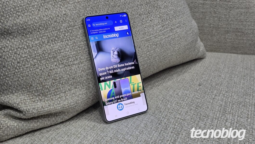Foto de um smartphone com tela acesa, posicionado verticalmente e levemente inclinado contra uma almofada de tecido cinza tramado. A tela exibe o site do Tecnoblog com a manchete "Dono de um DJI Romo hackeou quase 7 mil robôs aspiradores por acaso" e uma notícia sobre o Samsung Galaxy A57. No topo da tela, a URL "tecnoblog.net" e o horário "4:32". Há uma marca d'água branca do Tecnoblog no canto inferior direito, sobreposta ao fundo de tecido cinza que compõe o restante do cenário.