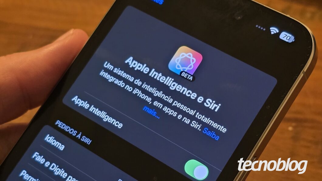 Um iPhone sendo segurado por uma mão exibe a tela de configurações da Apple Intelligence e Siri, com um design escuro. O texto informa que se trata de um sistema de inteligência pessoal integrado ao iPhone e à Siri, ainda em fase beta. O botão de ativação está ligado. A interface está em português, e no canto superior direito, há ícones de Wi-Fi e bateria indicando 70% de carga. O fundo da imagem tem uma superfície de madeira desfocada.