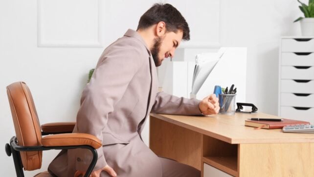 A man is sitting at a work desk holding his hip and grimacing as if in pain