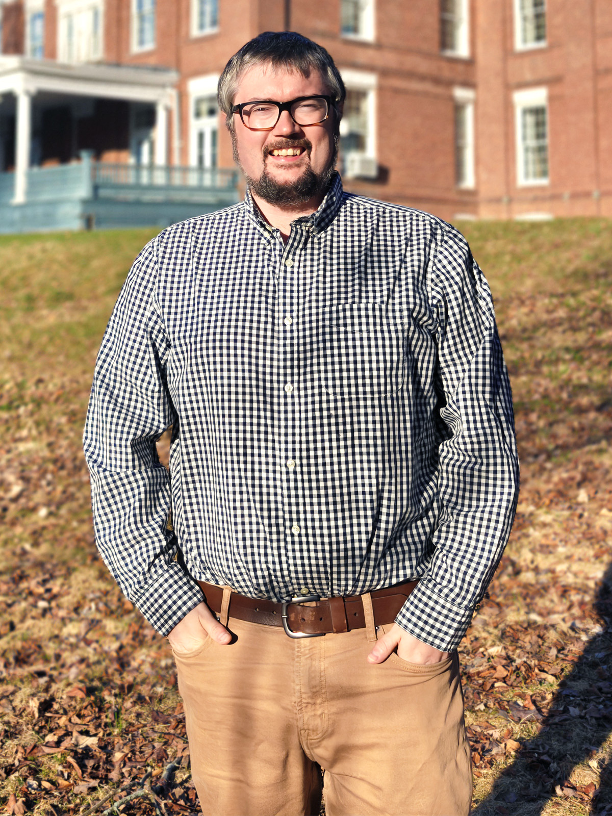 A man wearing a checkered shirt stands on the lawn in front of a brick building