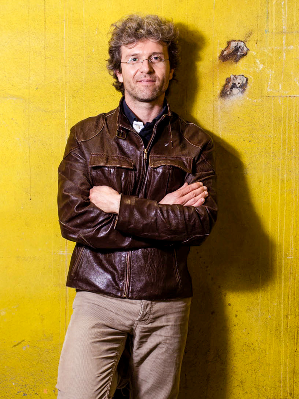 A man wearing a brown leather jacket leans against a neon-yellow wall.