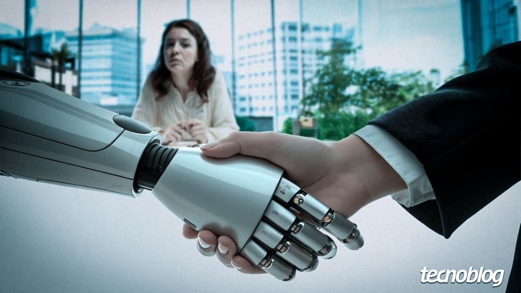 Em primeiro plano, um aperto de mãos entre uma mão robótica prateada, à esquerda, e uma mão humana, à direita. O robô possui dedos articulados com detalhes metálicos e pretos. O humano veste um paletó escuro. Ao fundo, uma mulher de cabelos castanhos e blusa clara está sentada à mesa, desfocada, observando a cena. O ambiente é um escritório moderno com janelas amplas e vista para prédios. No canto inferior direito, lê-se a logomarca "tecnoblog" em branco.