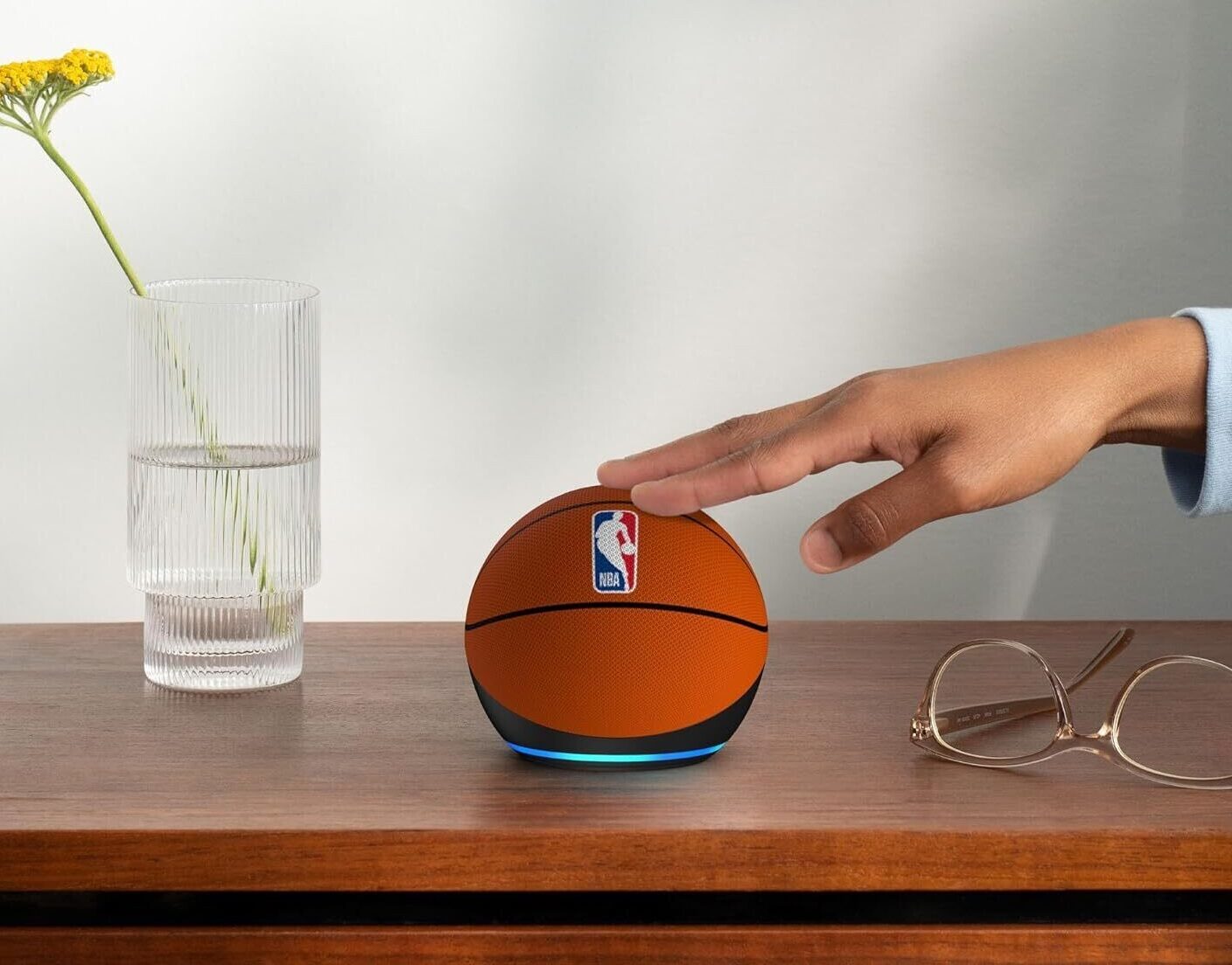 Echo Dot (5ª Geração) em edição limitada NBA, com formato esférico revestido em tecido laranja que imita a textura e as linhas pretas de uma bola de basquete. Na parte superior, destaca-se o logotipo oficial da "NBA" em azul, branco e vermelho. O dispositivo repousa sobre uma mesa de madeira escura. Uma mão de pele retinta toca o topo do aparelho. Ao lado, há um par de óculos de armação fina e um vaso de vidro canelado com flores amarelas, contra um fundo de parede cinza claro.