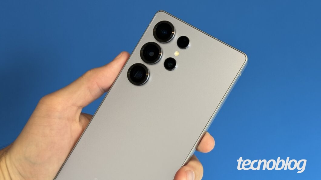 Uma mão segura um smartphone Samsung de cor prateada contra um fundo azul. O aparelho possui um design premium com cantos retos e uma configuração de câmera traseira composta por quatro lentes dispostas verticalmente no canto superior esquerdo. O logo da Samsung está gravado na parte inferior traseira do dispositivo.