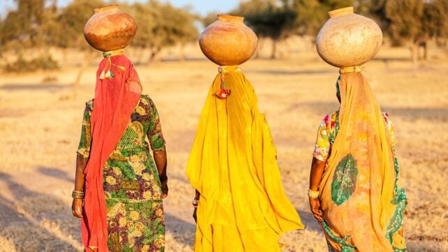 Three women in colorful clothing carry large water pots on their heads through a dry rural landscape in Rajasthan, India.
