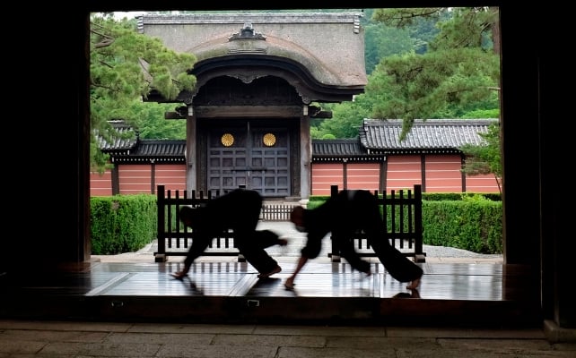 Two monks polish a wooden temple walkway, silhouetted against a traditional Japanese temple gate and garden.