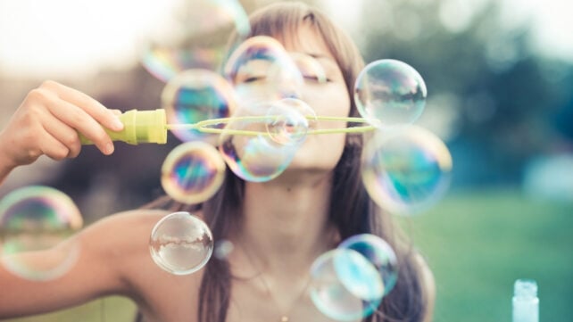A person outdoors blowing iridescent soap bubbles through a bubble wand.